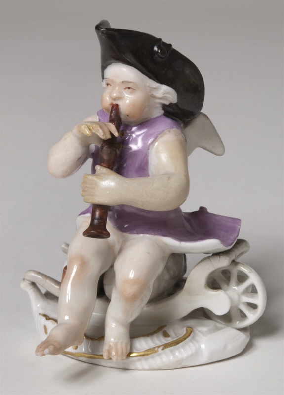 Figurin, bevingad putto sittande på lastad skottkärra blåsande flöjt